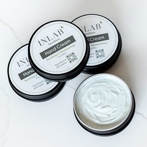 Four black containers of INLAB Soothing Hand Cream, with one lid removed to reveal the soft, rich cream inside, are beautifully displayed on a white marble surface.Four black containers of INLAB Soothing Hand Cream, with one lid removed to reveal the soft, rich cream inside, are beautifully displayed on a white marble surface.