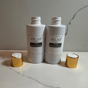 Two white cosmetic bottles labeled "INLAB" containing Cleansing Facial Gel Wash, with elegant gold caps, placed on a light-colored marble surface.
