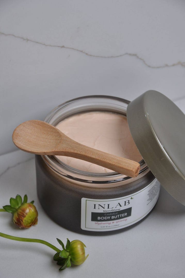 A jar of Emulsified Body Butter with a wooden spoon on top, partially open, showcasing its creamy texture against a marble background.